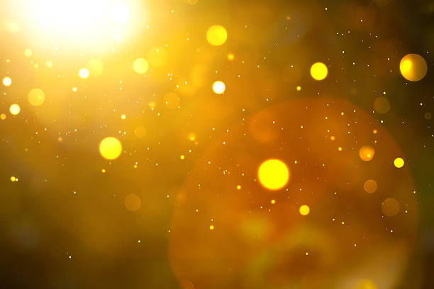 An abstract image showcases a rich golden bokeh effect, with varying sizes of blurred circular highlights creating a warm and luminous atmosphere. A bright lens flare emanates from the upper left, casting a radiant glow across the frame. Numerous tiny, sparkling particles are scattered throughout, adding a touch of magic and ethereal quality to the abstract composition.