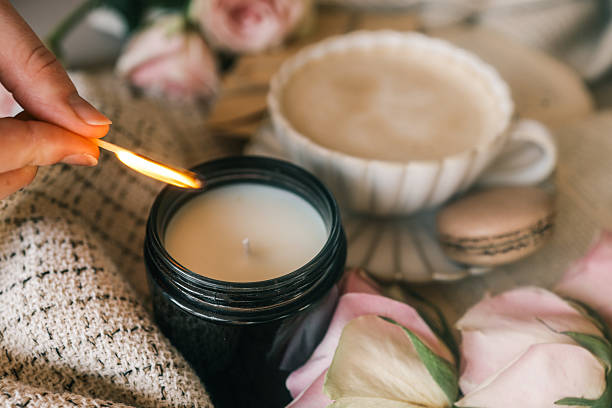 istock A cozy scene with a hand lighting a candle using a matchstick, surrounded by soft beige fabric, a cup of frothy coffee, pink roses, and a macaron. Warm and inviting atmosphere 2211217261