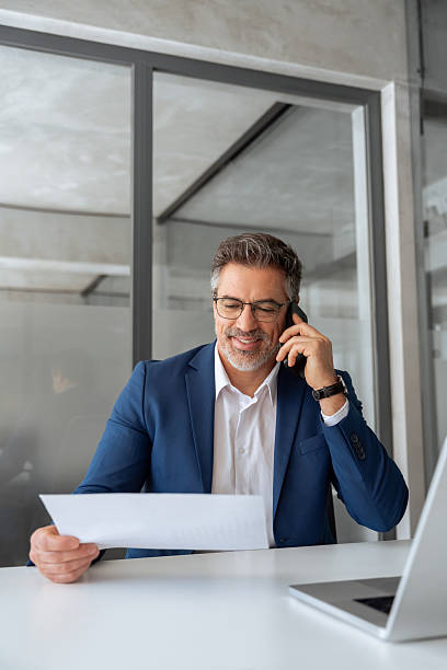 istock Positive financial specialist senior man working on laptop computer in office, having cell phone call. Mature businessman professional using pc, analysing business project, holding document. Vertical 2211221508