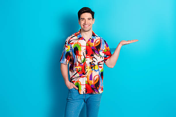 istock Smiling young man with colorful shirt poses against solid blue background, extending hand in presentation gesture 2211222524