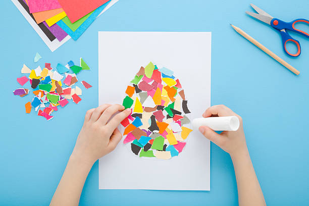istock Little child hands holding glue stick, gluing and making easter egg shape from colorful torn application paper pieces on white paper. Light blue table background. Pastel color. Closeup. Top down view. 2211225441