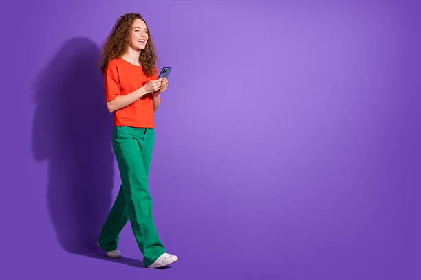 istock Young woman with curly hair using smartphone while walking against vibrant purple background in casual and stylish orange and green outfit. 2211225904