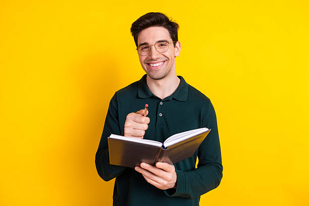 istock Handsome young man with glasses holding a notebook and pen, smiling confidently against a vibrant yellow background 2211227642