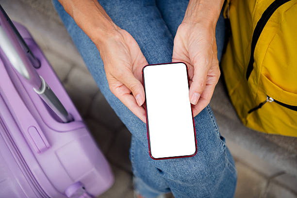 istock Mature woman holding smartphone displaying blank screen while traveling with suitcase outdoors during sunny day 2211230916