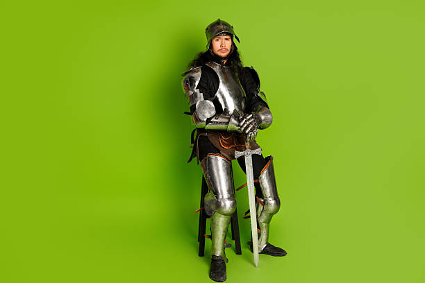 istock Medieval knight in armor with sword sitting on stool against vibrant green background 2211232657