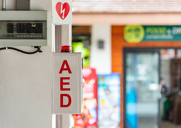 Front view of an automated external defibrillator (AED) unit in a red box installed in a recessed white wall cabinet, with instructional signage and symbols above it, photographed indoors for a clean medical safety concept.​The simple composition and clear labeling make this image ideal for themes related to emergency equipment, first aid, cardiac arrest response, health and safety regulations and public facility infrastructure.​