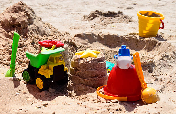 Children's plastic toys green end yellow car, shovel, yellow and red buckets, green ball with yellow sand on the beach by sea. Children's beach toys on sand on a sunny day. Sandbox on the playground
