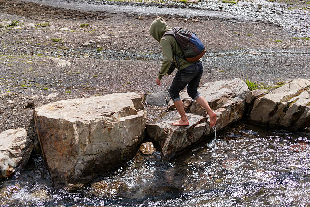 istock Barefoot man carefully balances on large rocks while crossing a shallow stream. Wearing a hoodie and backpack, navigating rugged terrain in an outdoor adventure setting, hiking in mountains 2211247024