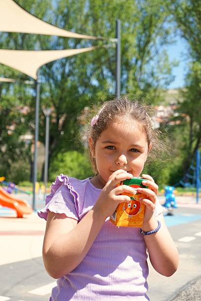 The child takes a break on the playground bench, juice in hand. Cheeks rosy from play, they watch others run and laugh. With every sip, they recharge—sweetness on their tongue, sun on their skin, joy all around.
