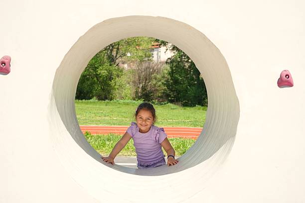 A little girl in a purple shirt smiles while posing through a circular opening of a modern playground structure. The background reveals green grass and trees, creating a cheerful and carefree atmosphere on a sunny summer day.