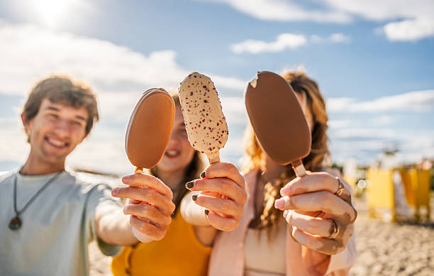 Three joyful friends enjoying ice cream bars on a sunny beach, soaking up the summer vibes and sharing laughter under the clear sky
