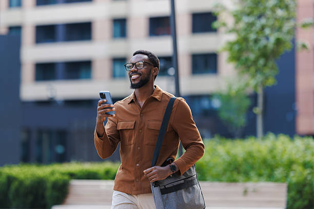 istock Smiling man walking outdoors while holding a smartphone and a shoulder bag 2211257182