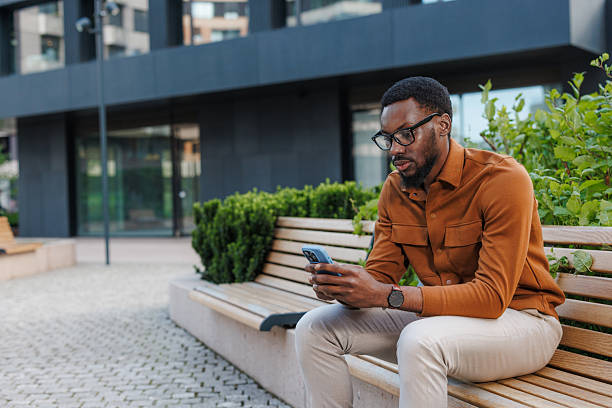 A Black man sitting on an outdoor bench, outside the office building, engrossed in using his smart phone. Urban greenery surrounds him, creating a relaxed yet focused atmosphere.