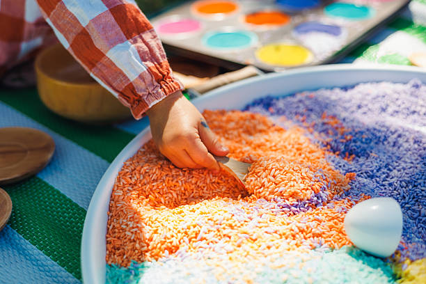 A hand reaches into a large tray filled with brightly colored rice during a sensory play session. This engaging activity takes place outdoors, fostering creativity and exploration among children.