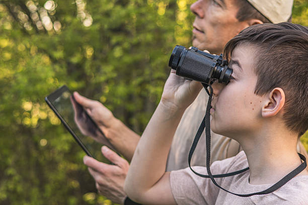 istock Birding. Father and son observing birds using binoculars and learning about them on digital tablet. Family time at weekend searching for birds or birdwatching in forest in late afternoon. 2211259481