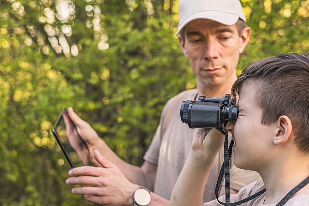 istock Birding. Father and son observing birds using binoculars and learning about them on digital tablet. Family time at weekend searching for birds or birdwatching in forest in late afternoon. 2211259492