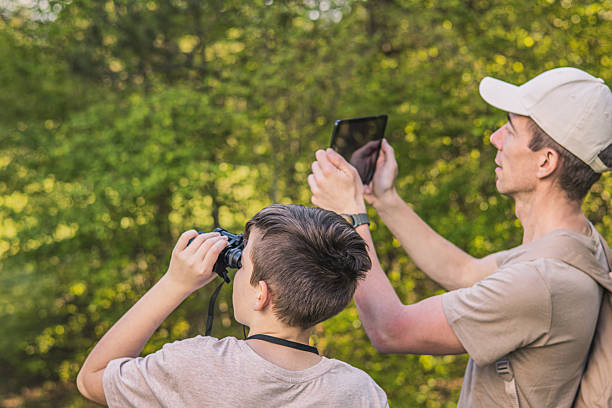 istock Birding. Father and son observing birds using binoculars and learning about them on digital tablet. Family time at weekend searching for birds or birdwatching in forest in late afternoon. 2211259556