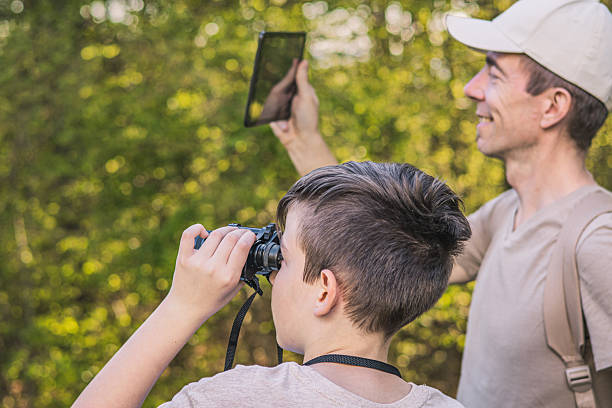 istock Birding. Father and son observing birds using binoculars and learning about them on digital tablet. Family time at weekend searching for birds or birdwatching in forest in late afternoon. 2211259574