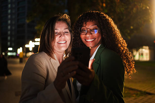 Two cheerful women enjoying together using a smartphone during the night. Joyful ladies sharing a delightful moment while using a mobile phone at dusk. Multiracial females having fun with a cellphone