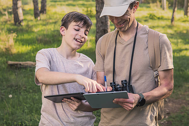 istock Birding. Father and son observing birds using binoculars and learning about them on digital tablet. Family time at weekend searching for birds or birdwatching in forest in late afternoon. 2211259659