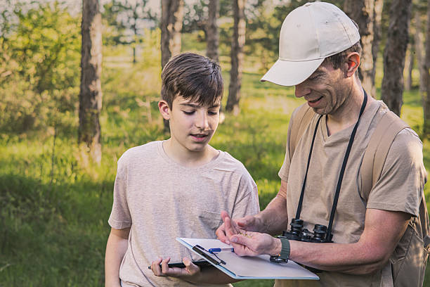 istock Birding. Father and son observing birds using binoculars, learning about them on digital tablet and feeding birds with birdseed. Family time at weekend searching for birds or birdwatching in forest in late afternoon. 2211259928