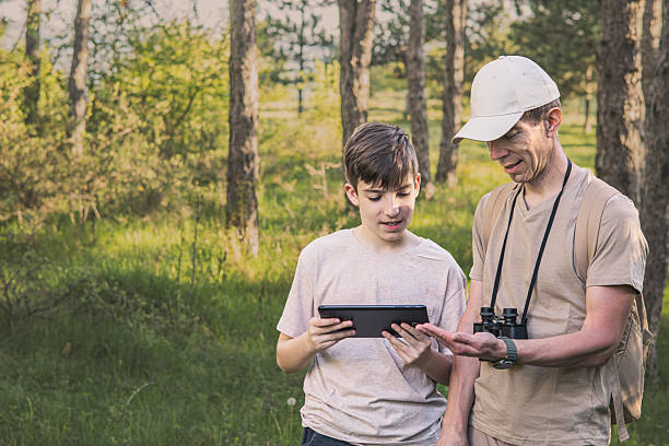 istock Birding. Father and son observing birds using binoculars, learning about them on digital tablet and feeding birds with birdseed. Family time at weekend searching for birds or birdwatching in forest in late afternoon. 2211260177