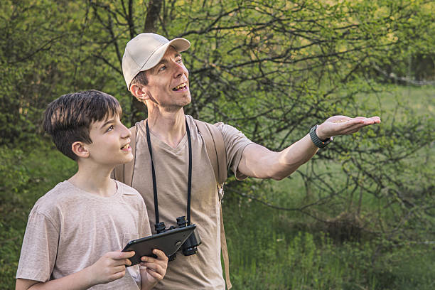 Birding. Father and son observing birds using binoculars, learning about them on digital tablet and feeding birds with birdseed. Family time at weekend searching for birds or birdwatching in forest in late afternoon.