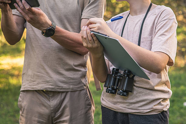 Birding. Father and son observing birds using binoculars and learning about them on digital tablet. Family time at weekend searching for birds or birdwatching in forest in late afternoon.