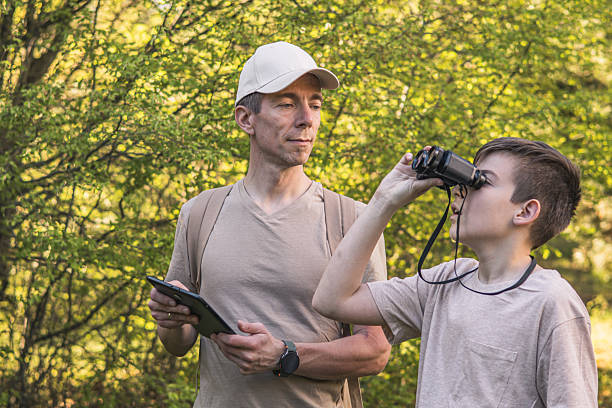 istock Birding. Father and son observing birds using binoculars and learning about them on digital tablet. Family time at weekend searching for birds or birdwatching in forest in late afternoon. 2211261487