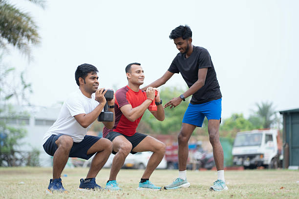 A fitness coach supervises two men performing squats with dumbbells during an outdoor training session. The participants, dressed in athletic gear, engage in strength training exercises on a grassy field surrounded by trees and natural elements. Emphasizing healthy lifestyle, teamwork, and outdoor fitness routines.
