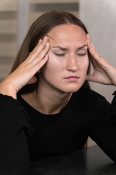 istock Young woman with closed eyes, touching her temples, experiencing intense discomfort from a severe headache while sitting at a table in her home, reflecting feelings of stress and fatigue 2211263257