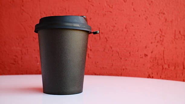 Black disposable coffee cup with lid on white table against vibrant red textured wall background