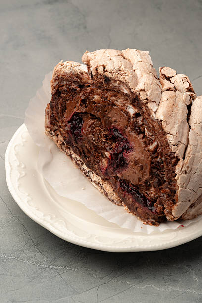 A chocolate meringue roulade is sliced and presented on a delicate white plate. The dessert features layers of rich chocolate and cherries, set against a simple gray background.