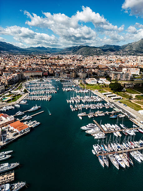 A picturesque aerial view overlooking the bustling harbor of Palermo, Italy. The scene showcases numerous yachts and boats docked, surrounded by the architectural charm of the city and scenic mountain views.