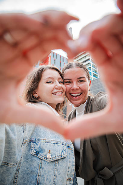 istock Vertical. Two cheerful young women posing together, creating a heart shape with their hands, symbolizing friendship, happiness, and love in an urban outdoor setting, sharing joy and positive emotions 2211287658