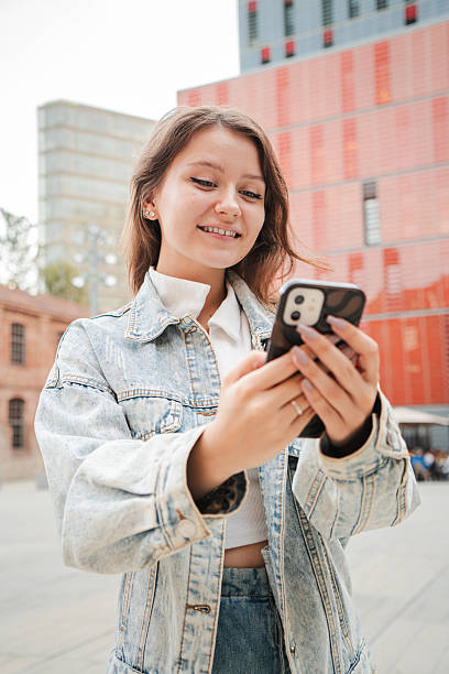 istock Vertical. Teenager girl smiling while using a cellphone outdoors, wearing a denim jacket, standing in front of red buildings and enjoying a casual moment while interacting with her smart phone. 2211287753