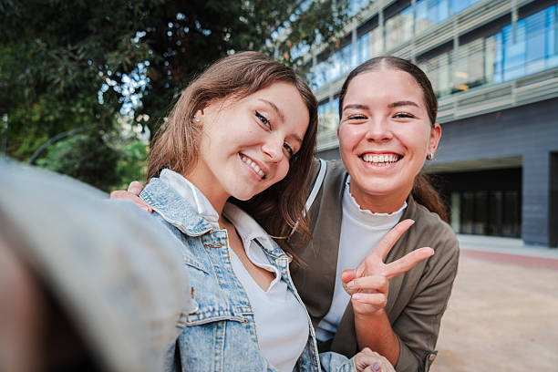 istock Two happy young women smiling and posing for a selfie outdoors, capturing a fun and joyful moment together while enjoying time outside in an urban setting, showing peace sign and friendship. 2211288092