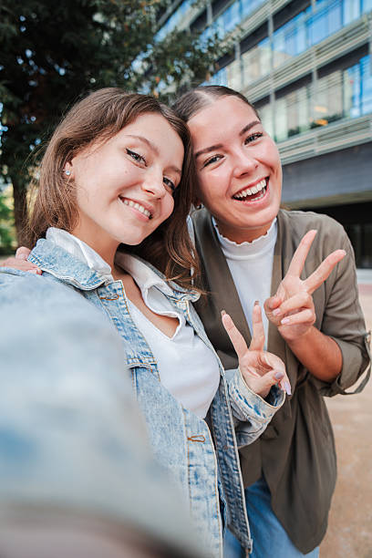 istock Vertical. Two happy young women smiling and posing for a selfie outdoors, capturing a fun and joyful moment together while enjoying time outside in an urban setting, showing peace sign and friendship 2211288322