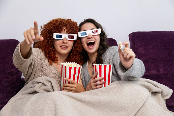 istock Two young women watching 3d movie with popcorn and blanket on sofa 2211290947