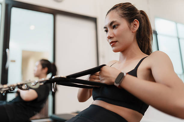 istock Focused Asian woman performing a resistance pull on a Pilates reformer, engaging her upper body and core workout in a modern studio. 2211303024