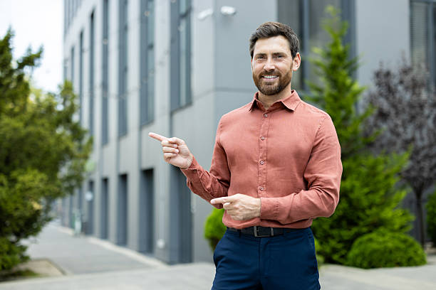 istock Cheerful businessman points to a modern building exterior while standing outdoors with a smile. 2211317060