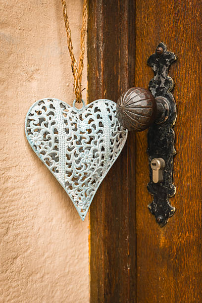 A beautifully crafted heart-shaped ornament dangles gracefully from a rustic door handle, accentuating the warm tones of an old wooden door in a cozy courtyard.