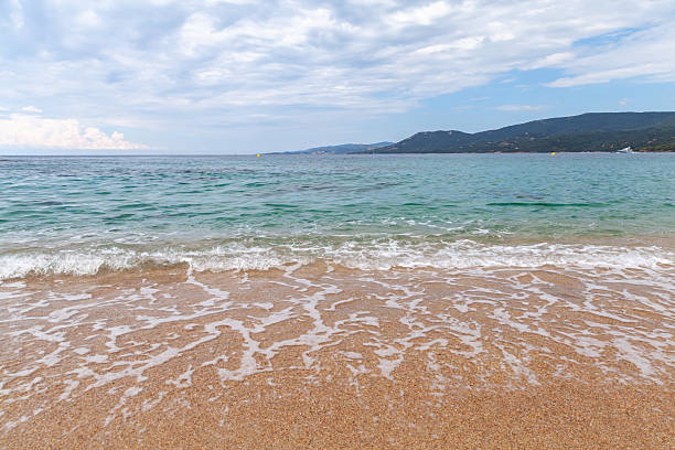 Tranquil beach scene featuring soft waves lapping sandy shores with hills in the distance under a partly cloudy sky. Perfect for concepts of relaxation, nature, and serene coastal escapes. Corsica