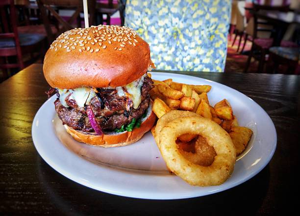 A colour photograph of a beef burger with chunky chips and onion rings on a round white plate.