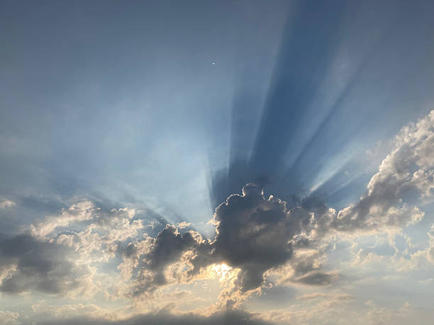 Image showcases a sunbeam phenomenon crepuscular rays during sunrise or sunset, with fluffy white clouds and soft pastel blue tones The photograph captures the interplay of light and clouds