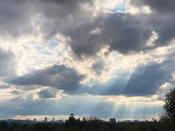A sunsetsunrise scene with dark gray clouds illuminated by the sun, contrasting against a cityscape below The image emphasizes light and cloud cover, creating a dramatic, moody atmosphere