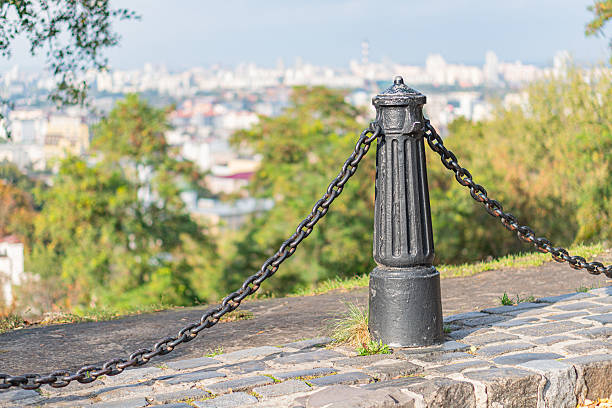 Image shows a historic landmark on a hillside, featuring a tall metal pole adorned with chains against a backdrop of cityscape and clouds Lush greenery covers the ground