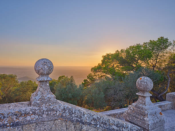 A serene stone terrace adorned with decorative columns, bathed in the warm hues of a sunset. The view overlooks the scenic landscapes of Majorca, Spain, featuring vibrant trees and a peaceful atmosphere.