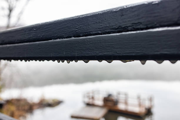 Raindrops delicately hang from a metal railing, with a blurred, misty waterfront and a wooden dock in the background, evoking a serene, tranquil feeling.