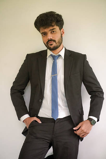 Stock photo shows a confident young adult businessman poses in a professional setting, dressed in a sharp suit, tie, and blazer. His poised demeanor and polished work attire reflect success, ambition, and modern professionalism